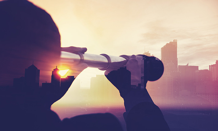Business man in suit with cityscape montage. The man is unrecognizable and you cannot see his face. He is superimposed onto a city skyline at sunset. He is holding a telescope looking into the city. Success, vision concept with copy space.